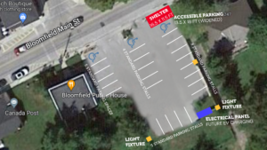 Image of the Bloomfield parking lot seen from overhead. The diagram shows the location of the bus shelter in the north eastern corner of the lot where an existing barrier curb will be removed. The ew lot design has a total of 22 parking spaces (9' wide), 3 accessible parking spaces (18' wide), three new overhead lights positioned at the back of the lot and along the eastern wall, plus an electrical panel in the back corner for future EV charging. 